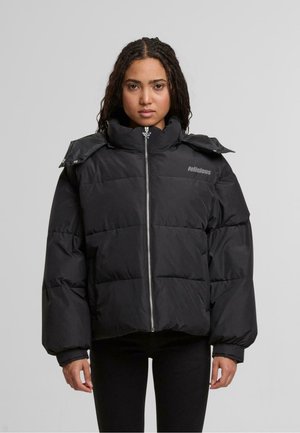 Mustapuffaparka, jossa on korkea kaulus, vetoketjukiinnitys ja kiiltävä pinta. Varustettu ylisuurilla, tikatuilla osilla ja logolla rinnassa.