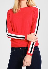 Pull en tricot rouge à manches longues avec des rayures noires et blanches. Encolure ronde classique, poignets côtelés et silhouette ajustée.