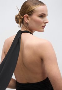 Rochie neagră cu gât halter, detaliu fluid în partea din spate, textură fină a țesăturii și talie cambrată. Modelul poartă cercei cerc și are o coafură îngrijită cu coc.