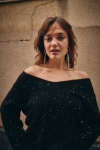 Moteris su pečius siekiančiais rudais plaukais dėvi juodą nuo pečių nukritusią megztinį su blizgučiais ir ilgus kabančius kristalinius auskarus priešais tekstūruotą sieną.