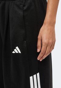 Gros plan d'une main reposant à côté d'un pantalon noir Adidas avec trois bandes blanches et le logo Adidas près de la poche.