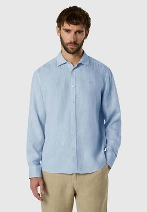 Uomo che indossa una camicia a maniche lunghe azzurro chiaro con bottoni e pantaloni beige, in piedi davanti a uno sfondo semplice grigio chiaro.