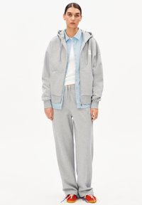 Grauer Kapuzenpullover mit Vorderentaschen, blaues Hemd mit Knöpfen, weißes Tanktop, graue Jogginghose und rote Schuhe mit mehrfarbigen Schnürsenkeln.