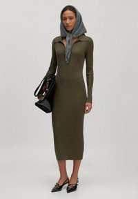 Vrouw in een aansluitende olijfgroene ribbed jurk, grijze hoofddoek onder haar kin vastgebonden, zwarte lakleren schoenen en een zwarte handtas in haar hand.