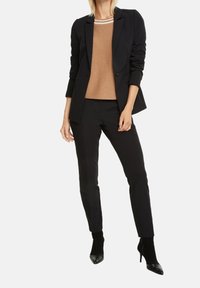 Blazer noir sur mesure porté sur un pull marron avec des accents rayés, associé à un pantalon noir fuselé et des escarpins noirs à bout pointu.