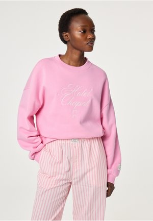 Vrouw draagt een roze sweatshirt met de tekst "Hotel Chapot" en roze-wit gestreepte wijde broek, staat met één hand in haar zak.