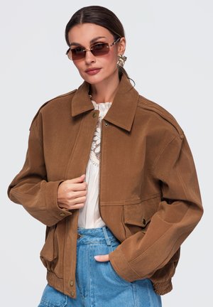 Femme portant une veste marron oversized, un haut en dentelle blanche, un jean bleu, de grandes boucles d'oreilles dorées et des lunettes de soleil sur un fond clair uni.