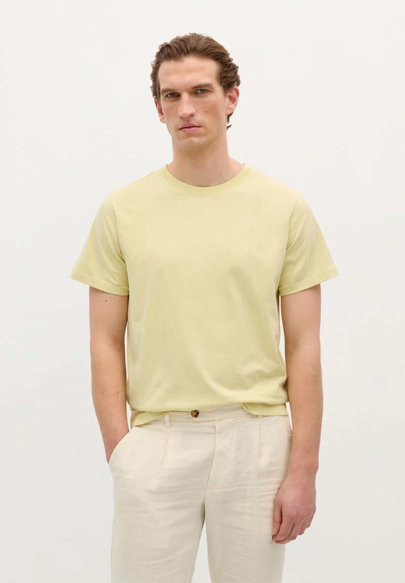 Lys gul bomulds T-shirt med korte ærmer og rund halsudskæring. Båret sammen med beige, plisserede linnedbukser. Enkel design, ingen mønstre.