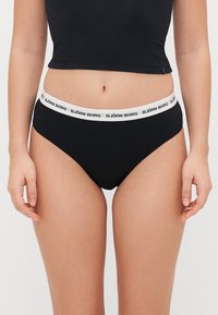 Schwarze Bikiniunterhose mit elastischem weißen Bund, der das Logo "BJÖRN BORG" trägt. Glatte Stoffqualität, niedriger Schnitt. Minimalistischer Stil.
