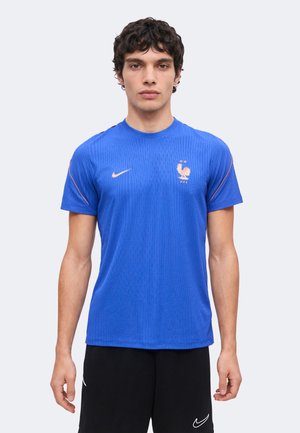 Jeune homme portant un maillot de football bleu de la France avec un emblème de coq doré et un short noir Nike sur un fond clair uni.