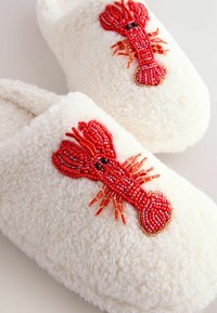 Weiße flauschige Hausschuhe mit rotem, verziertem Hummerdesign. Weicher Stoff, runde Form und dekorative Details auf der Vorderseite.