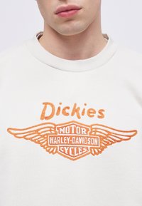 Gros plan d'une personne portant une chemise blanche avec un texte brodé orange "Dickies" et le logo Harley-Davidson Motor Cycles.