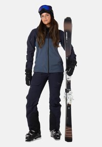 Skijakke i mørkegrå med sorte accenter, med lynlås foran. Båret med mørke bukser og sorte skistøvler, holder skis med hvide bindinger.