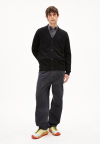 Schwarze Strickjacke mit Knöpfen, getragen über einem grauen Shirt. Kombiniert mit lockeren, dunklen Jeans und hellgelben Turnschuhen mit orangefarbenen Sohlen.