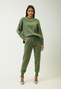 Groene gebreide trui met een ronde halslijn, versierd met decoratieve accenten, gecombineerd met bijpassende taps toelopende joggers. Sprankelende zilveren hakken.