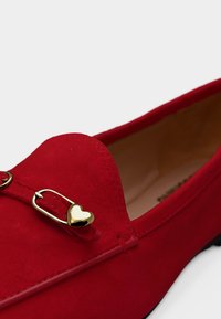 Κόκκινο μοκασίνι σουέτ με λεπτομέρεια από χρυσή αγκράφα σε σχήμα καρδιάς και μπεζ εσωτερική επένδυση σε λευκό φόντο.