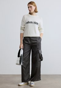 Biely pletený sveter s čiernym textom, v kombinácii s čiernymi saténovými širokými nohavicami s nákladnými vreckami. Modelka drží striebornú kabelku a má na sebe strieborné tenisky.