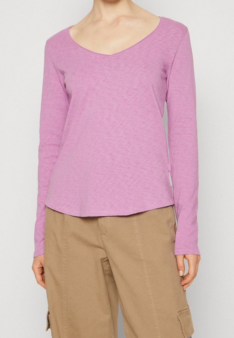 Pitkähihaisen V-kaula-aukon paidan vaaleanpunaisessa kankaassa, jossa on rento istuvuus ja hieman kaareva helma, yhdistettynä beigeihin cargo-housuihin.