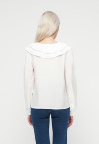 Hvid langærmet bluse med en plisseret flæsekant, letvægtsstof og en let tekstureret overflade. Båret med blå jeans.