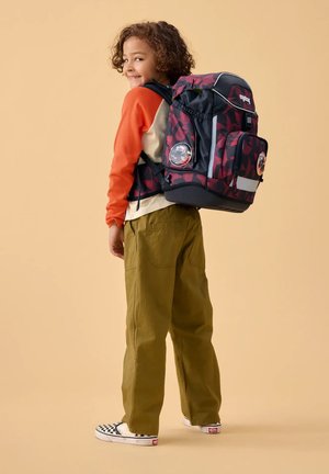 Smiling child with curly hair wearing orange and cream shirt, green pants, checkered slip-on shoes, carrying a large red and black backpack.