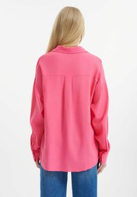 Košile na knoflíky z jasně růžové tkaniny, s límečkem, volným střihem a dlouhými rukávy s knoflíkovými manžetami. Zadní skládání dodává strukturu.