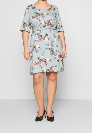 Nainen pukeutuneena vaaleansiniseen kukkakuviolliseen mekkoon, jossa on kyynärtaipeeseen ulottuvat hihat ja vyötärösolmittava nauha, yhdistettynä mustiin verkkokärkisiin terävälestisiin korkokenkiin.