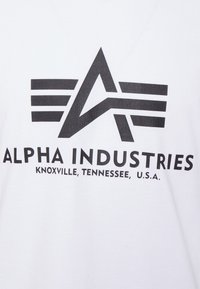 Bílá bavlněná tričko s velkým černým grafickým designem obsahujícím písmeno "A" a tři vodorovné pruhy nad textem "ALPHA INDUSTRIES".