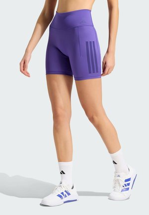 Persoon draagt paarse hooggetailleerde sportshorts, witte sokken met zwart logo en witte sneakers met blauwe strepen op een lichte achtergrond.