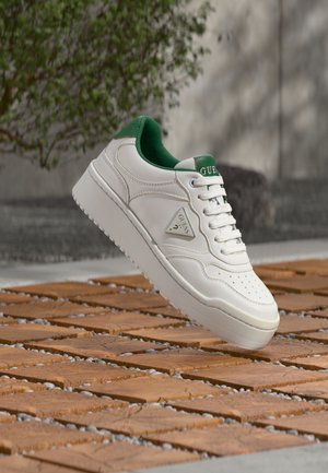 Baskets blanches avec doublure intérieure verte et logo "Guess" sur le côté, flottant au-dessus d'un sol extérieur en motifs bois et pierre.