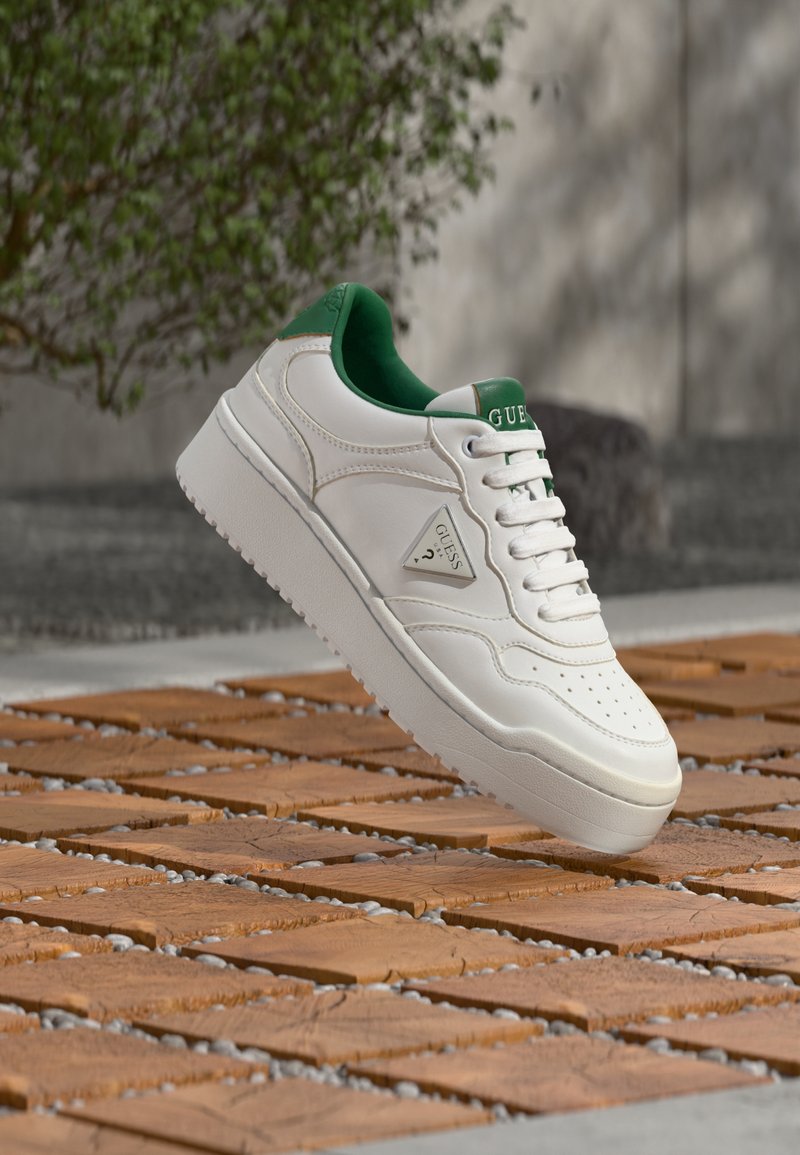 Baskets blanches avec doublure intérieure verte et logo "Guess" sur le côté, flottant au-dessus d'un sol extérieur en motifs bois et pierre.