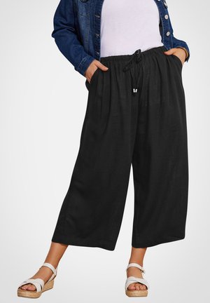 Person iført sorte, vidde bukser med snor, hvide sandaler, hvid top og blå denimjakke, hænder i lommerne, stående.