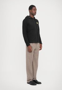 Black zip-up hoodie with a gold logo, paired with light brown textured trousers and black slip-on shoes. Simple design and neutral colors.