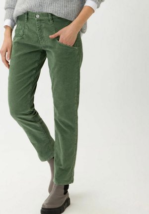 Frau trägt grüne Cordhosen, grauen Pullover und graue Stiefeletten, eine Hand in der Tasche, steht vor weißem Hintergrund.