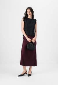 Ženska stoji ob beli ozadju, oblečena v črno vezeno bluzo in maroon krilo, drži majhno črno torbico.