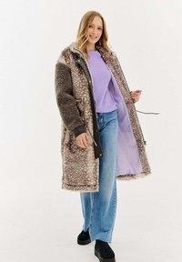 Žena v dlouhém hnědém kabátu s tečkami z umělé kožešiny, pod kterým má fialový svetr a modré džíny, se usmívá a drží kabát otevřený.