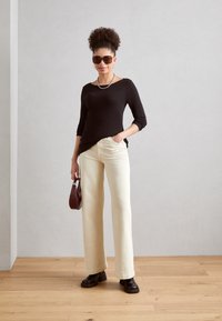 Haut noir à manches longues, jean large crème, chaussures à talons marron et lunettes de soleil. Les accessoires incluent un petit sac à main marron et un collier en argent.