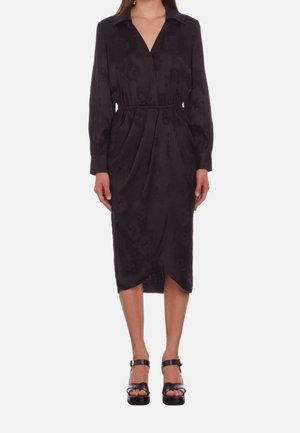 Frau trägt ein langärmliges schwarzes Midikleid mit Blumenmuster, tailliert, mit Kragen und überlappendem Saum vorne, kombiniert mit schwarzen Plateau-Sandalen.