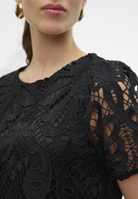 Haut noir avec un design en dentelle intricé, présentant des motifs texturés sur les épaules. Le tissu lisse sous la dentelle met en valeur le détail et le contraste.