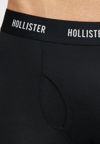 Κοντινό πλάνο στη ζώνη μαύρων μπόξερ με την επιγραφή «Hollister», που φοριέται πάνω σε γυμνό δέρμα.