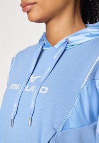 Gaiši zils Mizuno hoody ar auklu kapuci, kuram ir balts logs, gluda tekstūra un kontrastējoša plecu paneļa dizains.