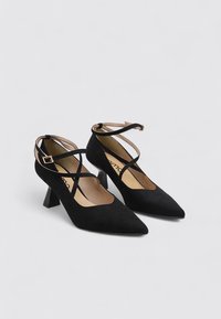 Zapatos de tacón alto de ante negro con puntera afilada, que presentan correas cruzadas en el tobillo y un tacón ancho para mayor estabilidad. Forro interior suave.