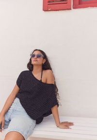Maglione nero in maglia oversize con scollatura asimmetrica, abbinato a pantaloni corti di denim chiaro. La modella indossa occhiali da sole a forma di gatto e si sieda su una panchina.