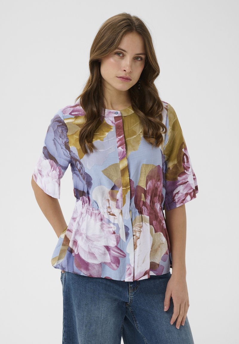 Žena s vlnitými hnedými vlasmi má na sebe viacfarebnú kvetovanú blúzku s tromi štvrťovými rukávmi a modré džínsy, stojaca pred jednoliatym pozadím.