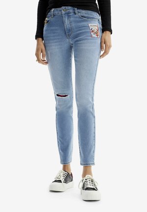 Person trägt hellblaue Skinny-Jeans mit einem abgenutzten Knie und einem bunten Aufnäher am Oberschenkel, kombiniert mit schwarzen und weißen Sneakers.