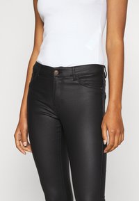 Pantalons slim noirs en matière lisse et extensible, dotés d'une fermeture à bouton sur le devant et de poches latérales avec des accents de couture discrets.