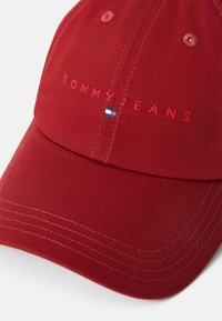 Rød bomuldsbat med broderet "TOMMY JEANS" logo, der har blå og hvide flag accenter. Struktur design med syet detalje og buet skygge.