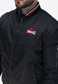 Czarna kurtka bomberka wykonana z gładkiego materiału, z czerwonym i białym ściągaczem, haftowanym logo oraz bocznymi kieszeniami z zamkiem.