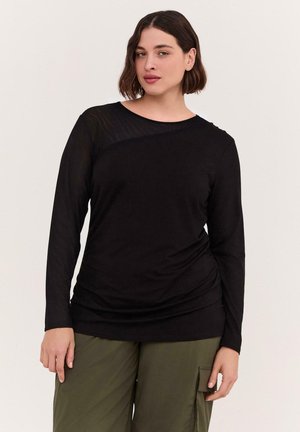 Kvinde med kort brunt hår iført en sort langærmet top og olivengrønne cargobukser mod en ensfarvet lys baggrund.