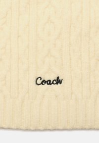 Kremowa dziana tkanina z warkoczowymi wzorami i czarnym haftowanym logo "Coach" umieszczonym centralnie w pobliżu dolnej krawędzi.