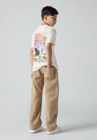 Ανοιχτόχρωμο κρεμ μπλουζάκι με πολύχρωμο τυπωμένο σχέδιο στην πλάτη, συνδυασμένο με μπεζ παντελόνι φάρδους και λευκά αθλητικά παπούτσια.